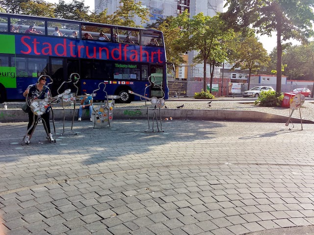 The Beatles Monument in Hamburg. Five metal cut-outs holding instruments. A woman is posing into one of the cut-outs.